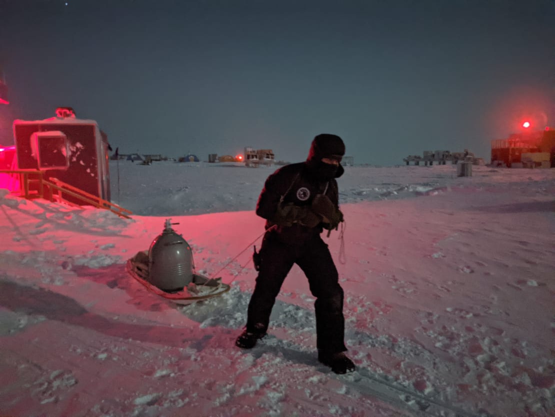 Person dragging large metal container on a sled across the snow.