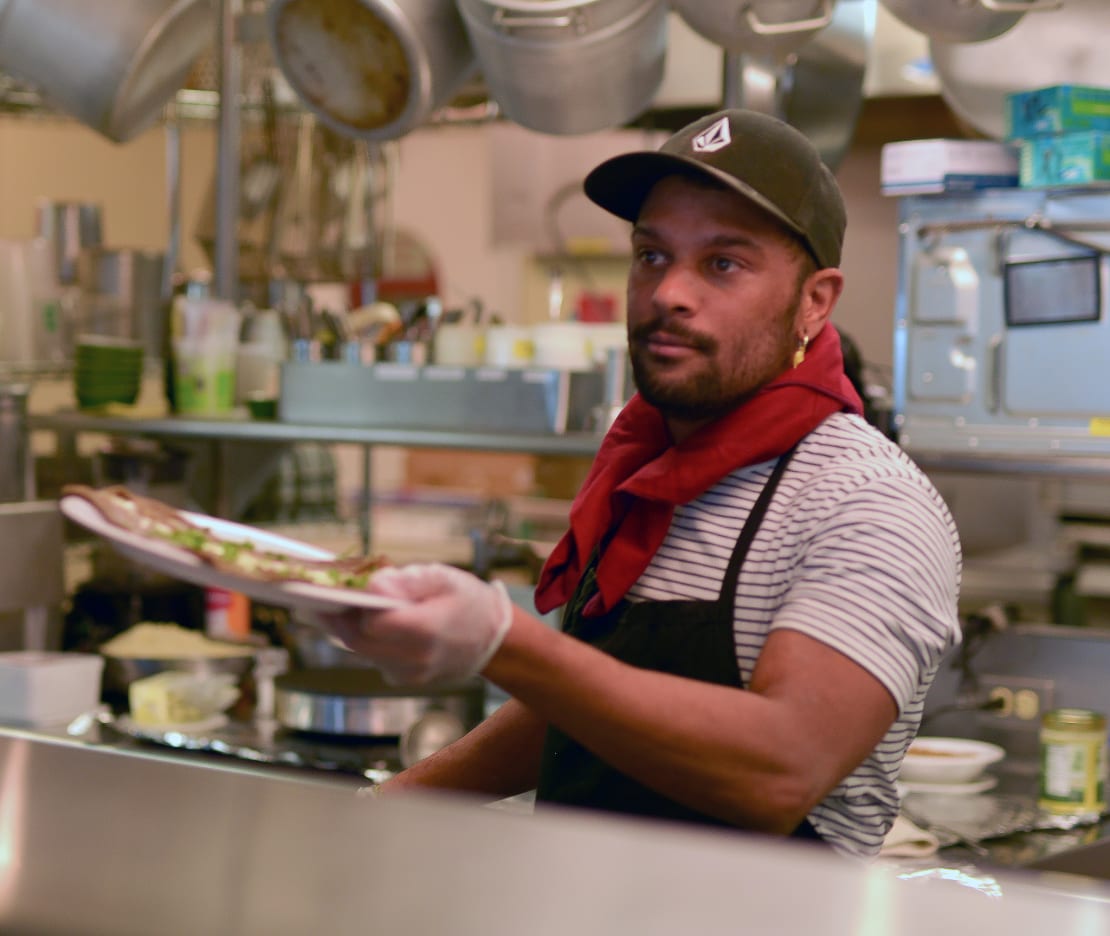 Winterover Celas in South Pole kitchen, handing off a plated crepe to someone off camera.