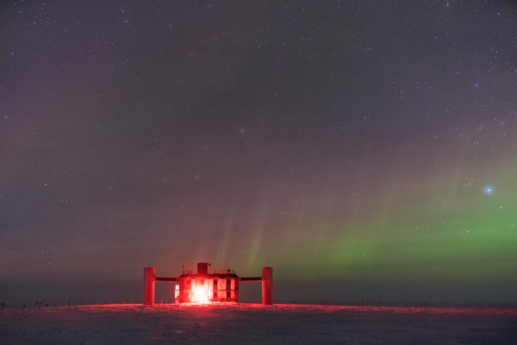 IceCube Lab lit up in red lights under starry sky with low faint auroras.