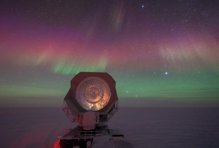 View of the South Pole Telescope dish with skies full of green and purple auroras.