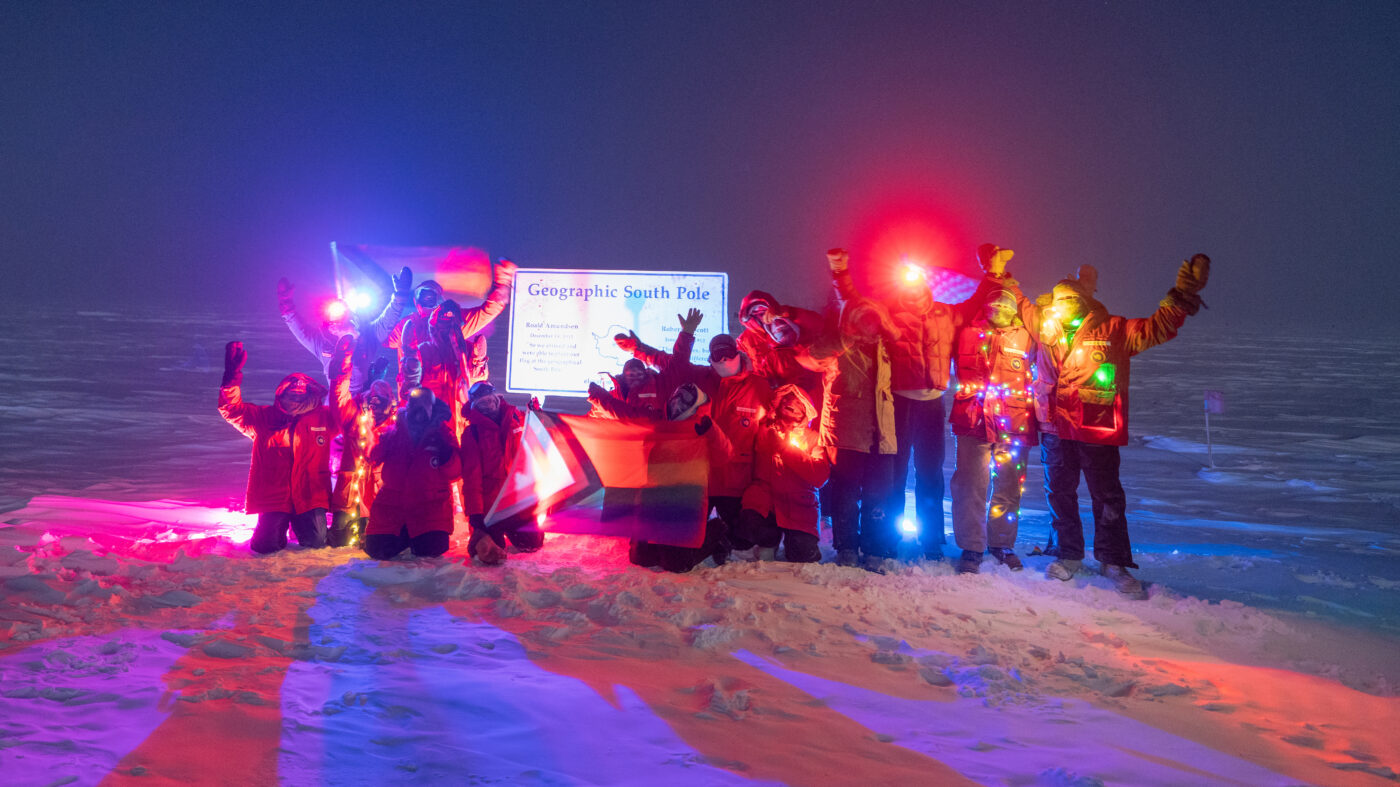 Group photo with Pride flag in front of geographic South Pole sign.