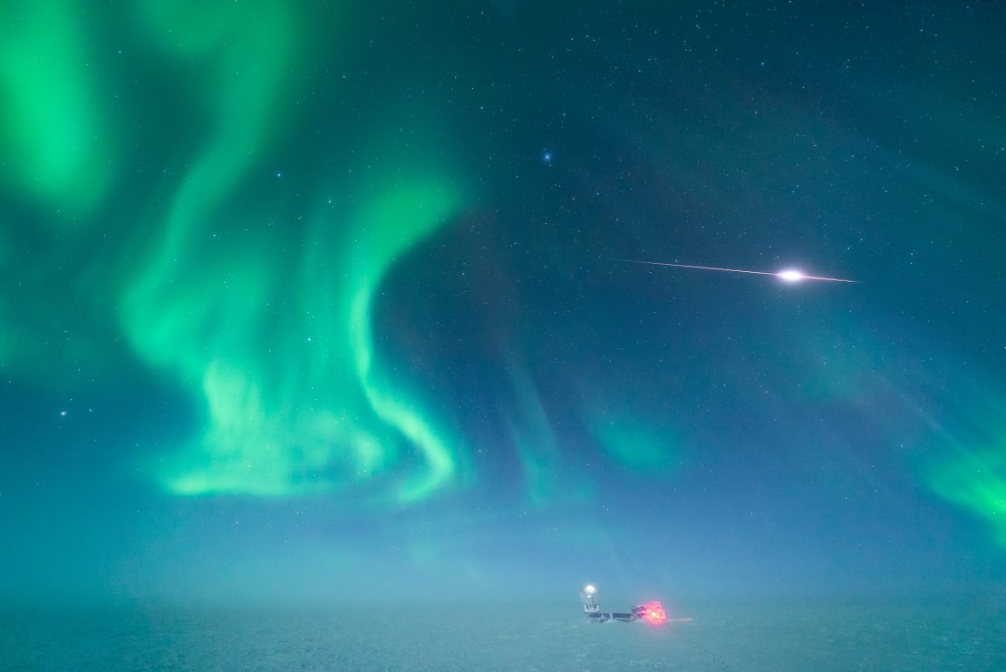 A meteor streaking through an aurora-filled night sky over the South Pole Telescope in the distance.