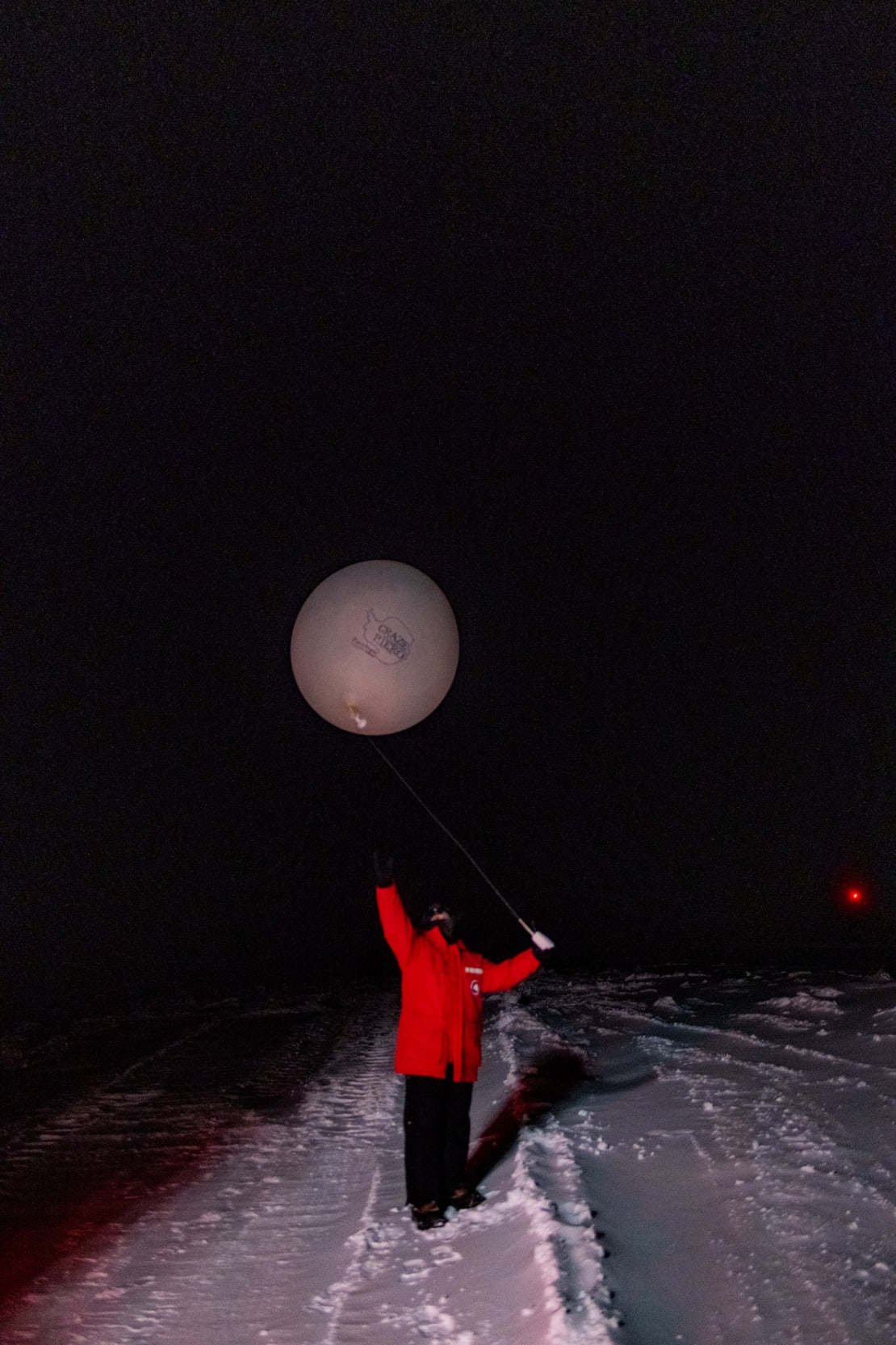 Winterover in red parka launching a radio balloon outdoors in darkness.