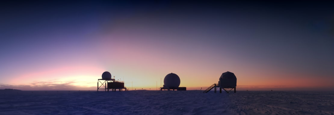 Colorful sunrise at the South Pole with three satellite domes in silhouette in the distance.