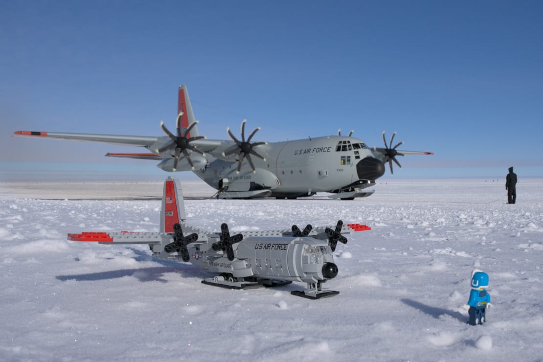 An LC-130 plane parked on the ice and a person facing it, and a small LEGO replica of the scence on the ice in the foreground.