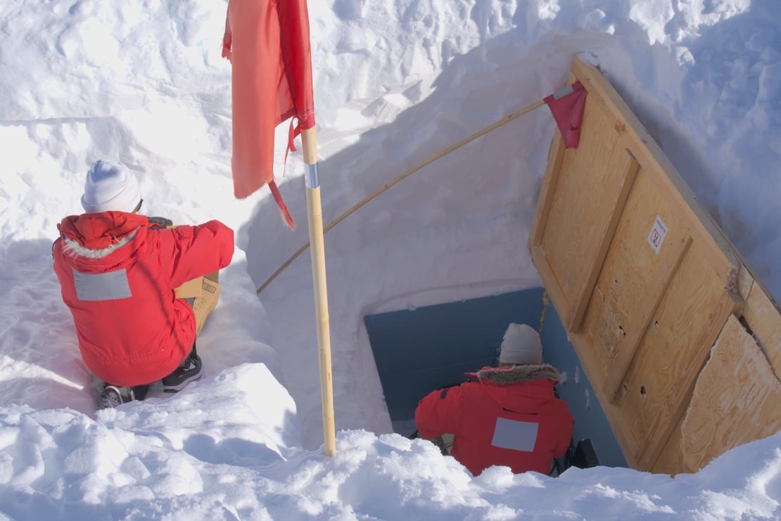 Two people in red parkas working outside at the South Pole in an open vault that was dug out from under the snow and ice.