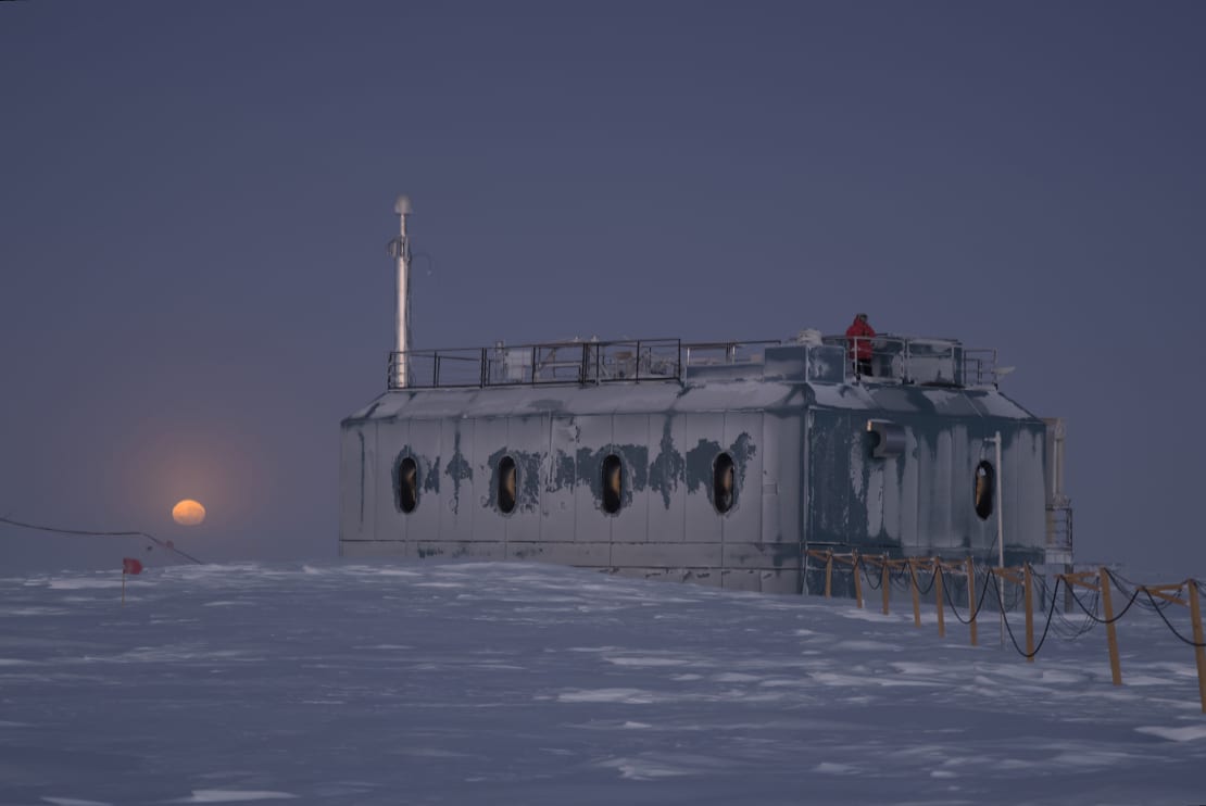 Moon low on horizon during sunset at the South Pole, a frosty building in the foreground, sky and ground all bluish in hue.