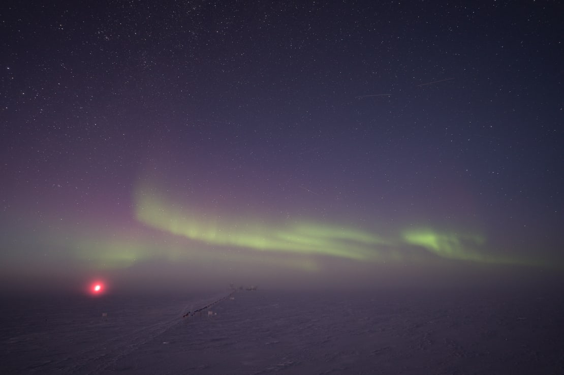 A line of low auroras seen over horizon of the Dark Sector at the South Pole, with red light in distance, which is the IceCube Lab.