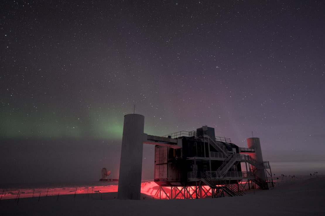 The IceCube Lab in shadow, red lights seen below the structure, faint auroras and stars in sky overhead.