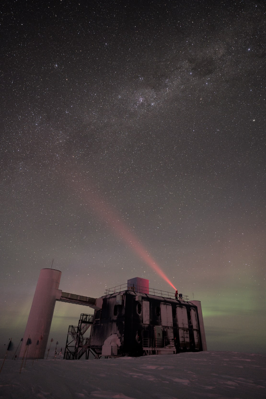 Starry sky above the IceCube Lab at the South Pole, with a winterover on the rooftop shining headlamp light into the sky.