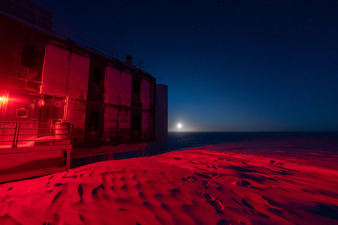 Moon just over horizon against a dark but bluish sky, with IceCube Lab lit in red to the left in the foreground.