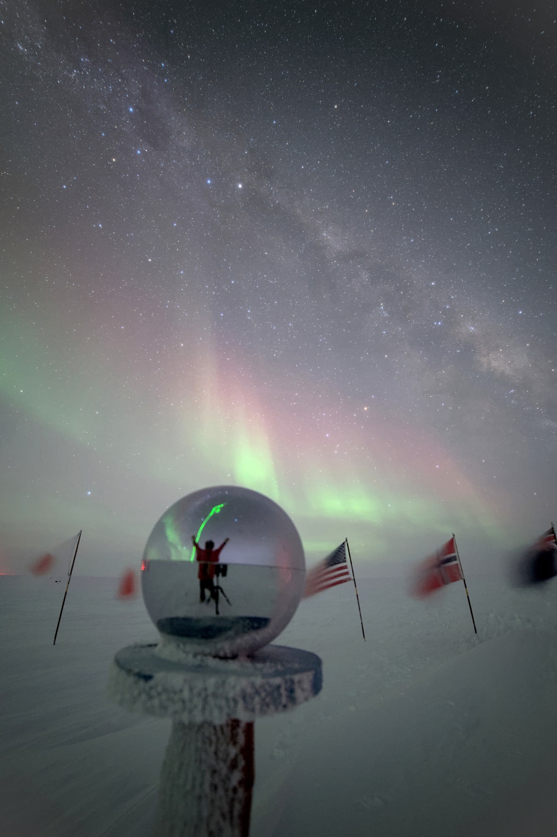 Reflection of winterover with arms up overhead seen in the mirrored globe marker at the ceremonial South Pole.