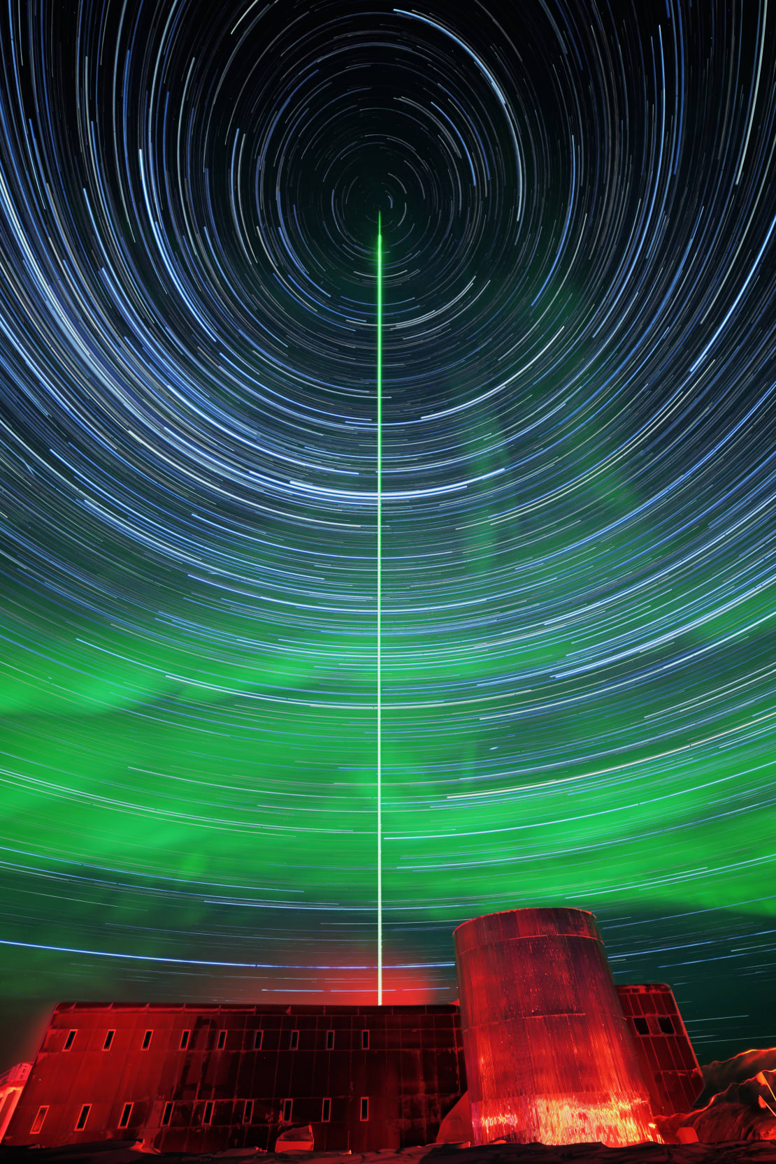 Blue lines of concentric circles from a three-hour exposure fill the night sky, with the South Pole station lit in red at the bottom and with green auroras just above the station.