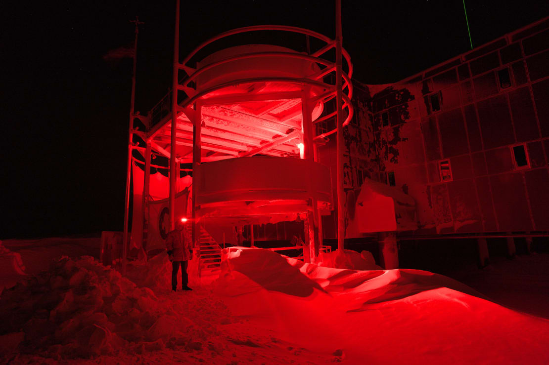 A winterover standing in the dark, under red light, near a somewhat tall snowdrift at the station entrance.