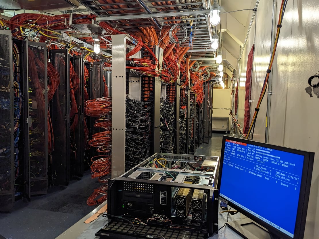 Indoor view of monitor and computer in foreground with rows of racks and orange cables in background.