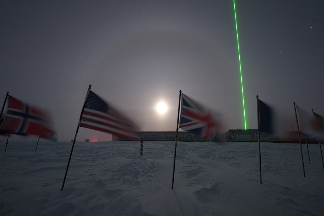 Bright moon with halo seen between flags at ceremonial South Pole, a bright green laser directed up into the sky from the station in the background.