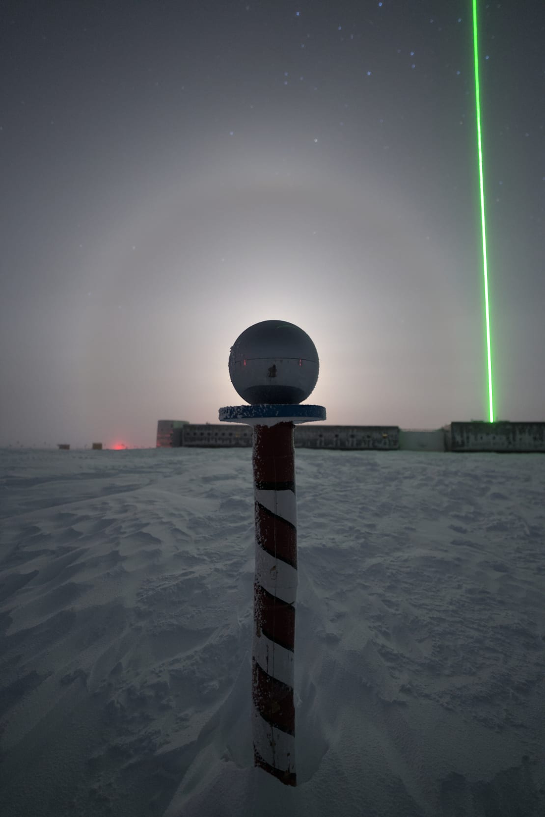 Bright moon with halo hidden behind spherical marker at ceremonial South Pole, a bright green laser directed up into the sky from the station in the background.