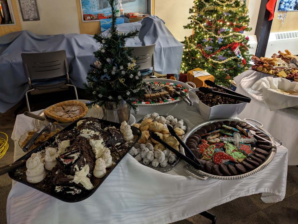 A table loaded with trays of cookies and treats, decorated Christmas tree in background.