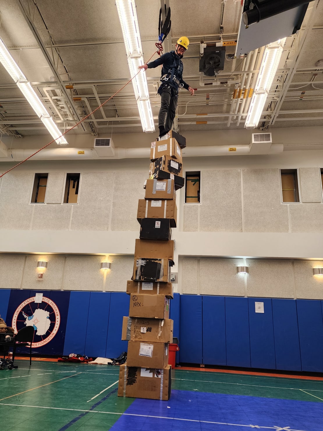 Person attached to ceiling-hung harness standing on top of tower of cardboard boxes, arms out to sides.