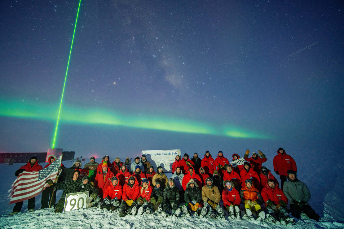 South Pole winterover group photo under a star-filled bluish sky with a horizontal streak of green auroras and a vertical green laser beam shooting up into the sky.