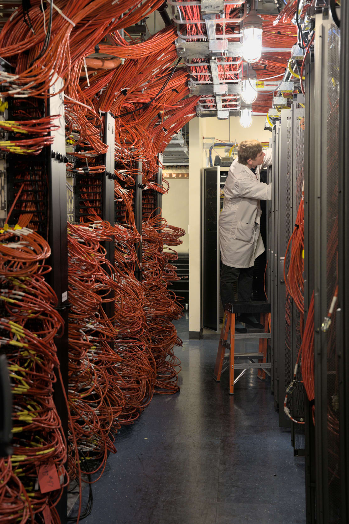 Winterover standing on stepladder in server room with floor to ceiling bundles of orange cables.