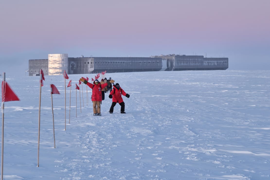 Several people in red parkas, some waving to camera, along flagline as they walk back to South Pole station.