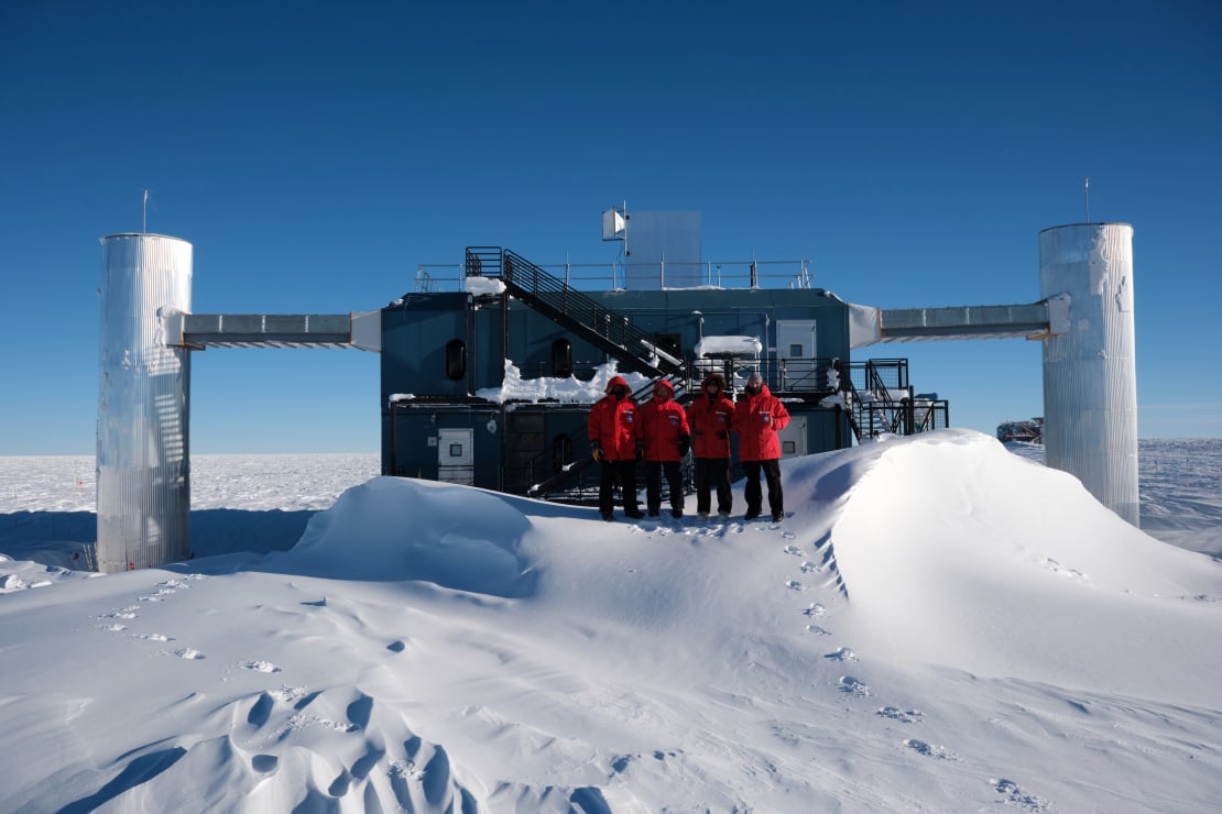 Four winterovers in red parkas posing on a snow drift in front of the IceCube Lab on bright sunny day.