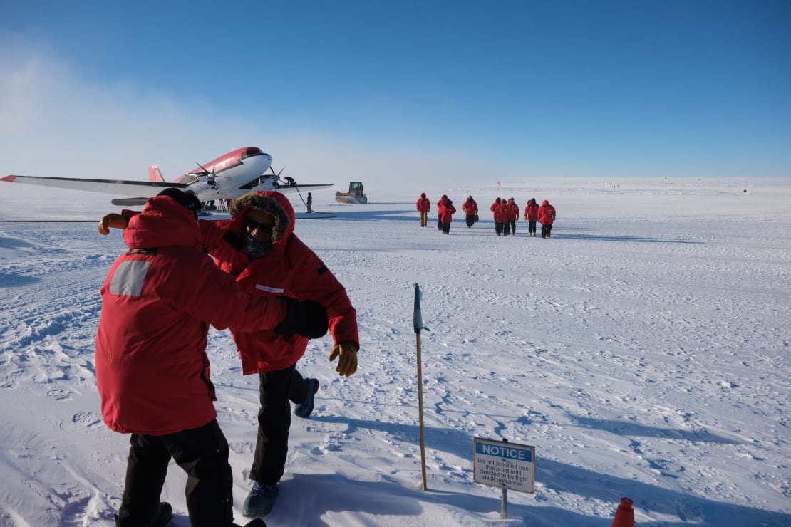 Plane parked on ice in background with group of arrivals nearby, two people in red parkas approaching for a hug in the foreground.