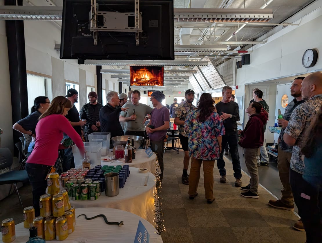 Group of people mingling over cocktail hour inside galley of South Pole station, ceiling monitors displaying cozy fireplace image.