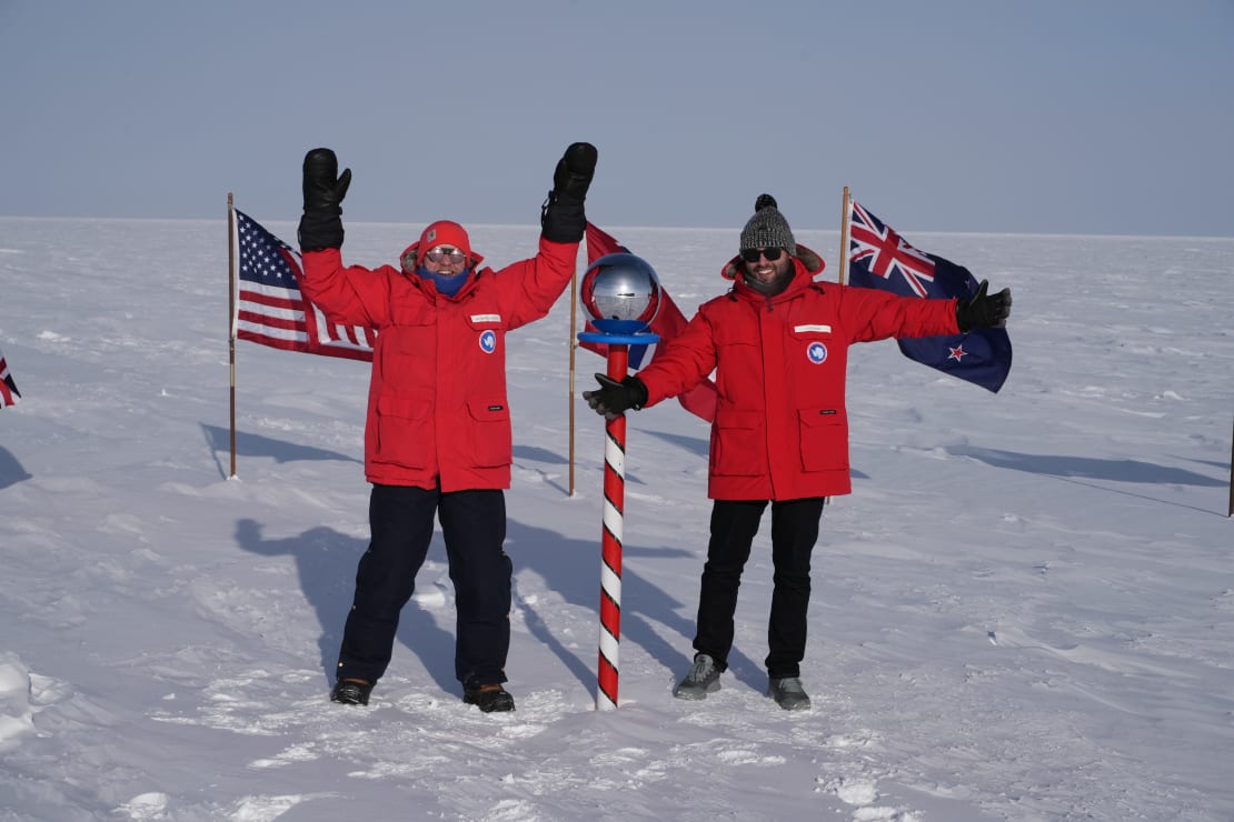 Two men in red parkas standing at the ceremonial South Pole